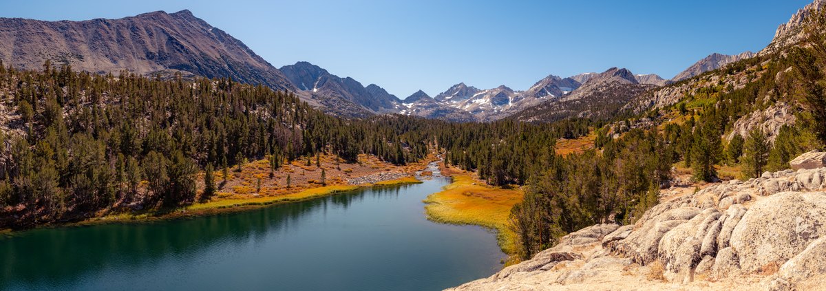 Golden autumn grasses trace the edge of a clear mountain lake beneath granite peaks and a wide blue sky.