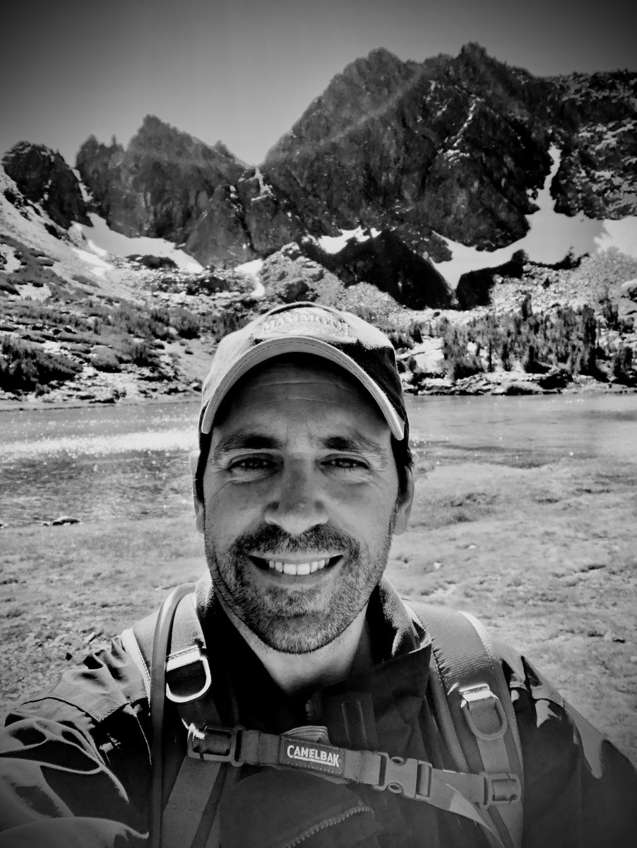A black-and-white self-portrait of James Casalino at an alpine lake with rugged peaks rising behind him.