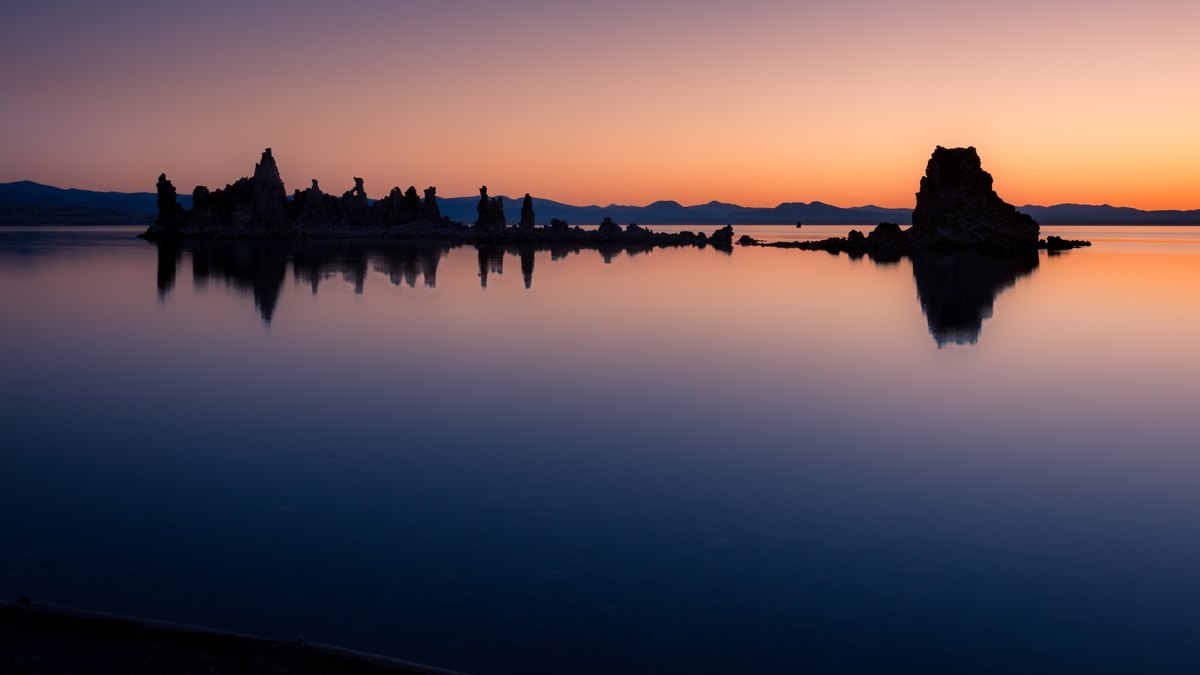 Pastel dawn light spreads across calm water as a line of tufa towers and distant mountains repeats in reflection.