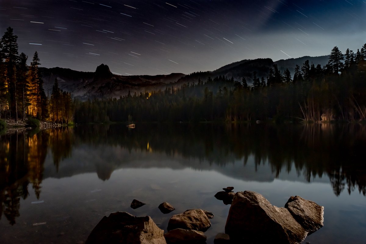 Star trails sweep over a forested mountain lake, where warm shoreline lights shimmer in the dark water.